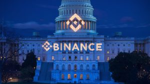 A photo of the Binance logo projected onto the US Capitol building at night. The logo is large and bright, with white text that says "Binance" in large, blue letters. The building is illuminated, with its columns and dome casting interesting shadows. The background contains trees and a few buildings. The overall scene has a blue hue.