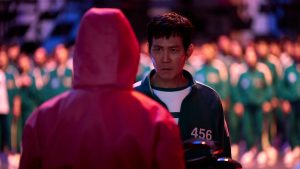 Man in pink hoodie with back to the camera, facing a man in Squid Game with green jacket on. Masses of people in the background.