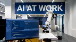 A photo of an office space with a computer monitor displaying a coding interface. There's a large sign above the monitor that says 'AI at work'. There's a robotic arm in the background. The robotic arm is holding a pen and is writing on a piece of paper. The office space has a modern design with white walls and a grey floor.