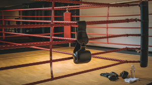 A photo of a boxing ring with red ropes and a black leather belt hanging on one of the ropes. The ring is in a gym with a wooden floor. There are gloves, socks, and a water bottle near the ring.